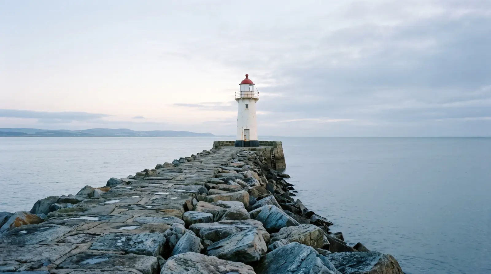 A small white lighthouse at the end of a stone breakwater at dawn, calm grey sea