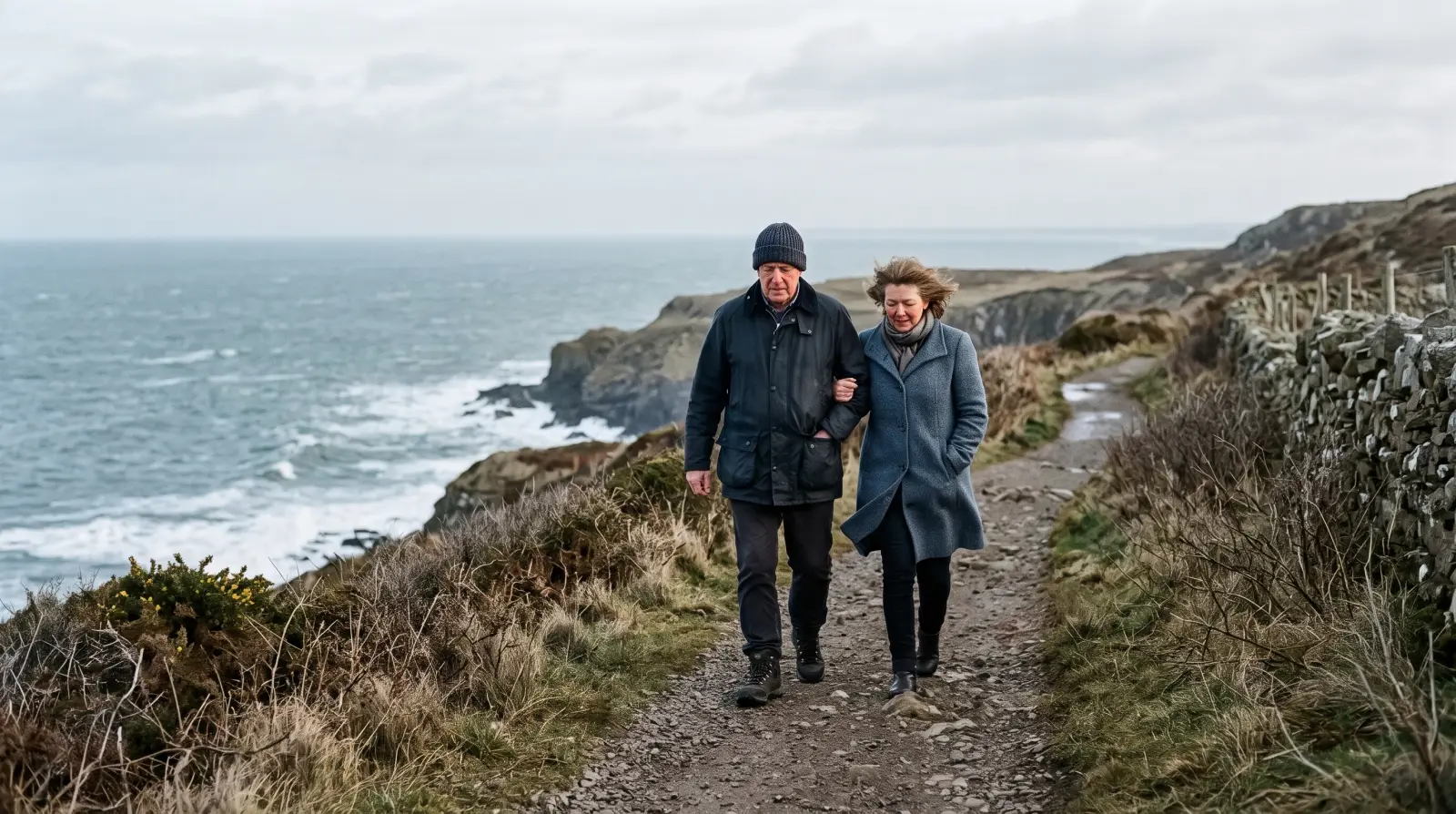 A couple in their sixties walking arm in arm along a coastal path on a cold, bright morning, the sea beside them