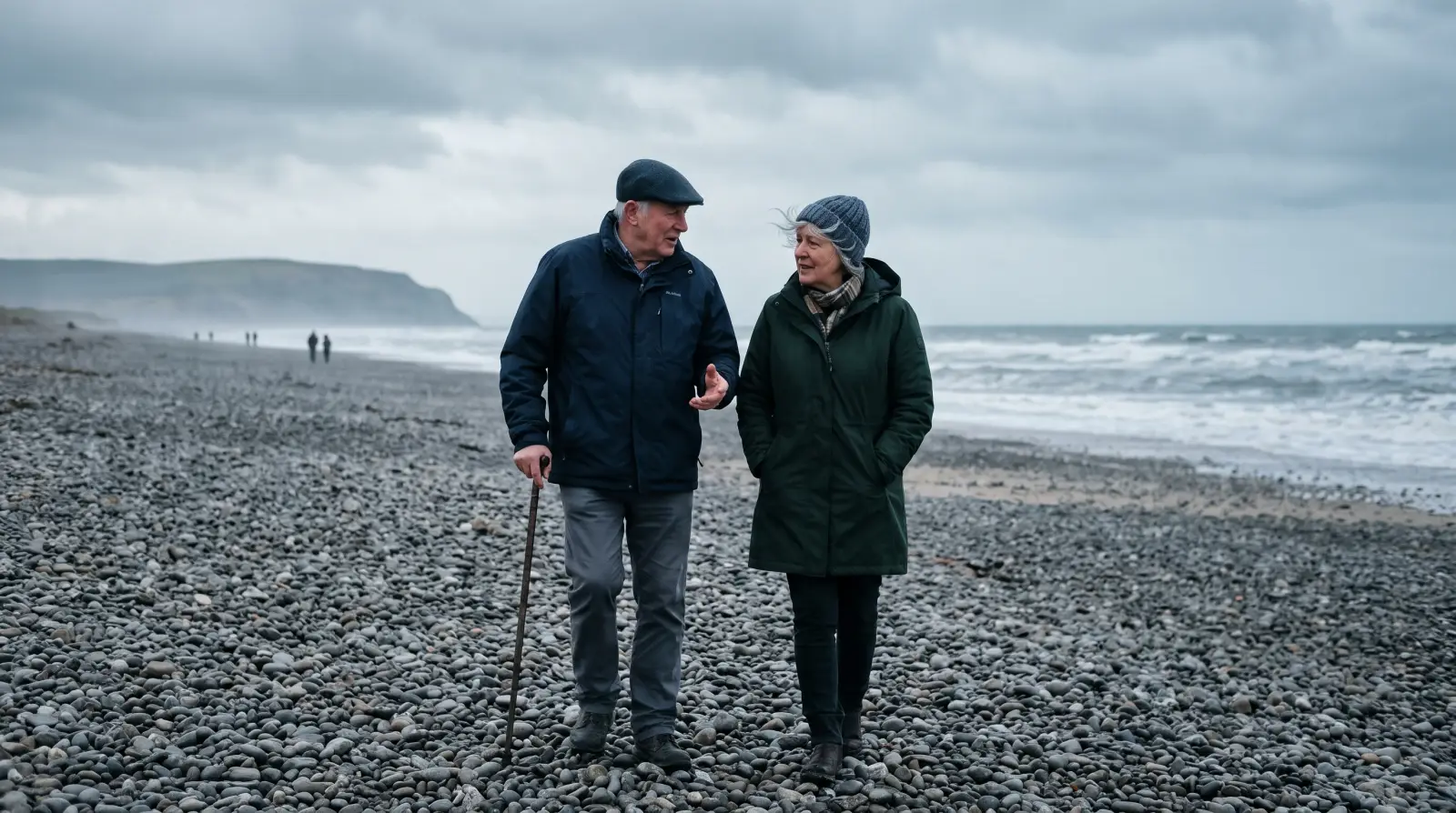 A couple in their sixties in winter coats walking and talking along a shingle beach under an overcast sky